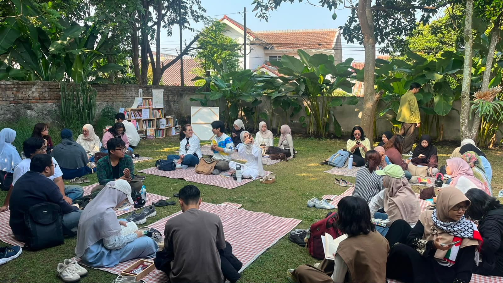 Puluhan orang duduk lesehan di atas tikar di taman, membaca buku bersama dalam kegiatan Bagi Baca.